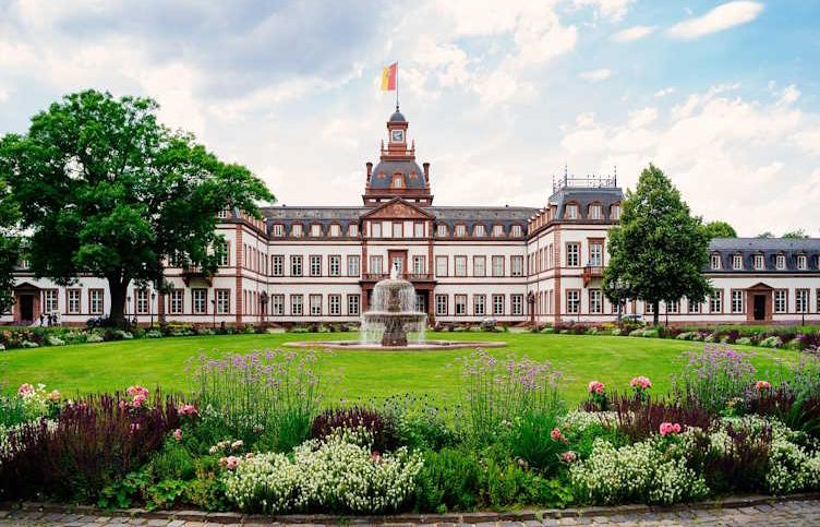 Schloss Philippsruhe in Hanau mit Garten und Statue im Vordergrund, umgeben von Bäumen und Blumen.