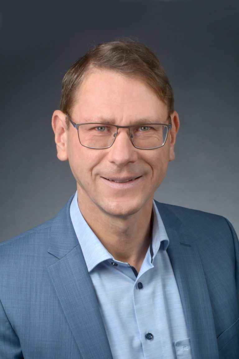Mann mit Brille und Anzughemden, lächelt freundlich vor grauem Hintergrund.