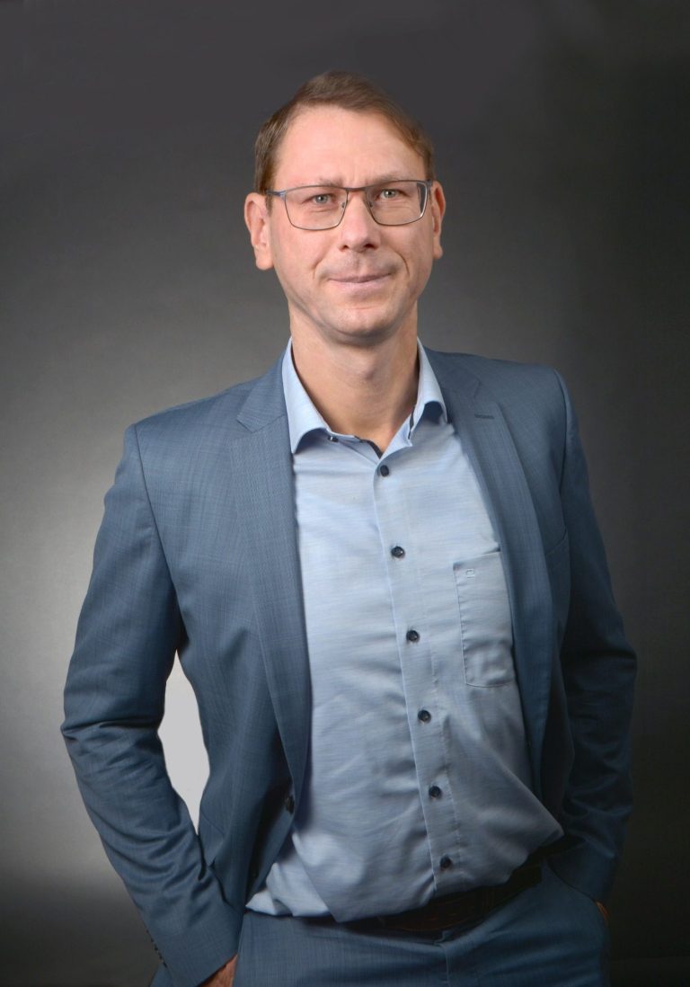 Mann in Anzug mit Brille, steht in lässiger Pose vor neutralem Hintergrund.